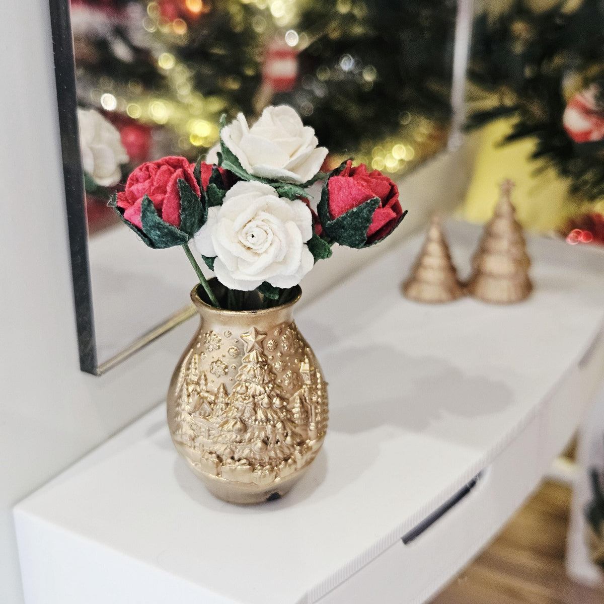 Wider view of red and white roses in a gold-coloured Christmas tree vase on a white sideboard, with festive decorations behind.