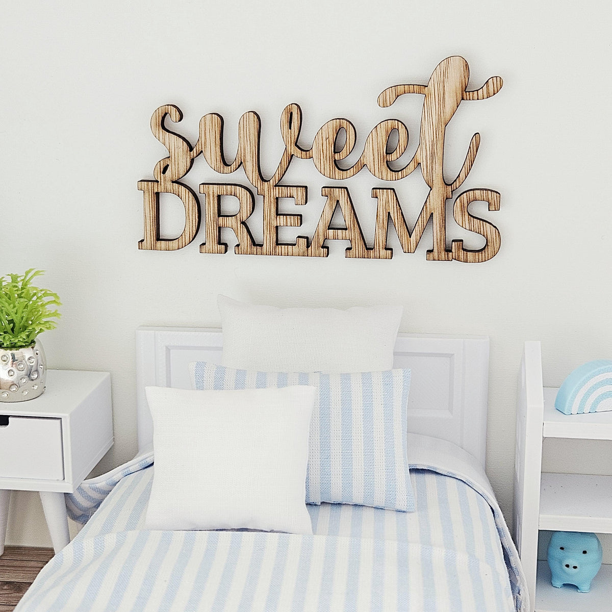Sweet Dreams sign displayed above a blue-striped miniature bed, with a potted plant to the left and white shelving to the right.