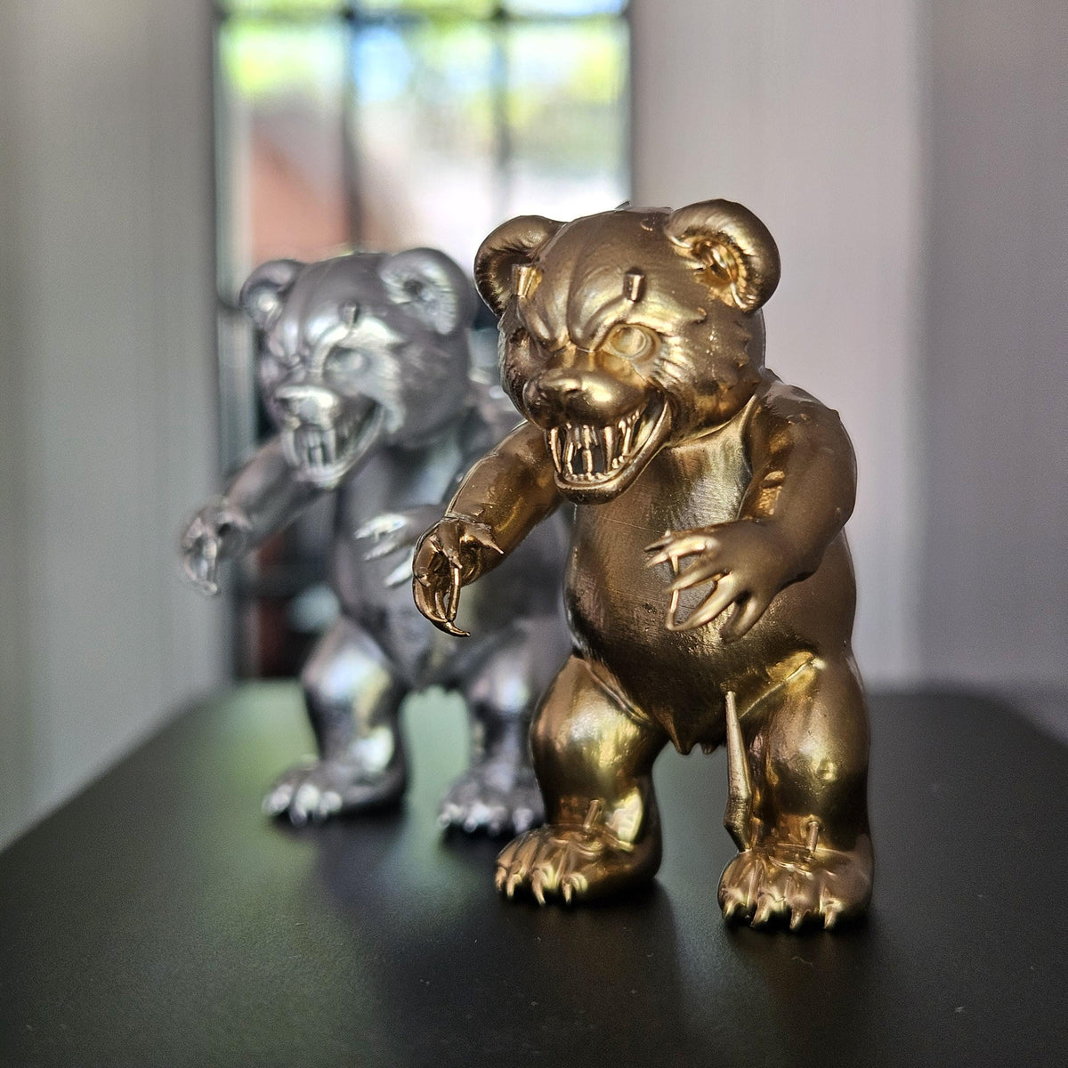 Three-quarter close-up of the gold scary bear ornament, showing bared teeth and extended claws, with the silver version behind.