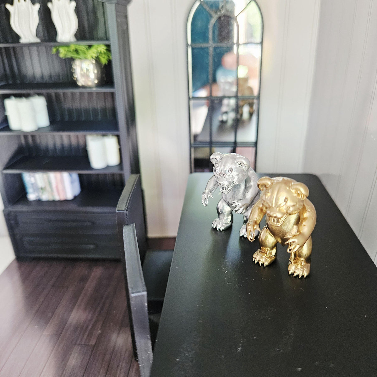 Elevated view of silver and gold scary bear ornaments on a black table in a miniature room with shelving and an arched mirror.