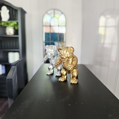 Gold scary bear ornament with a silver version behind it, displayed on a black table beside a miniature chair.