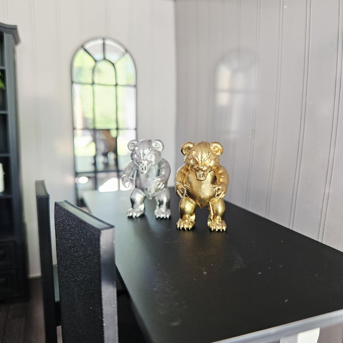 Wide view of silver and gold scary bear ornaments on a black table, with a miniature chair and arched mirror nearby.