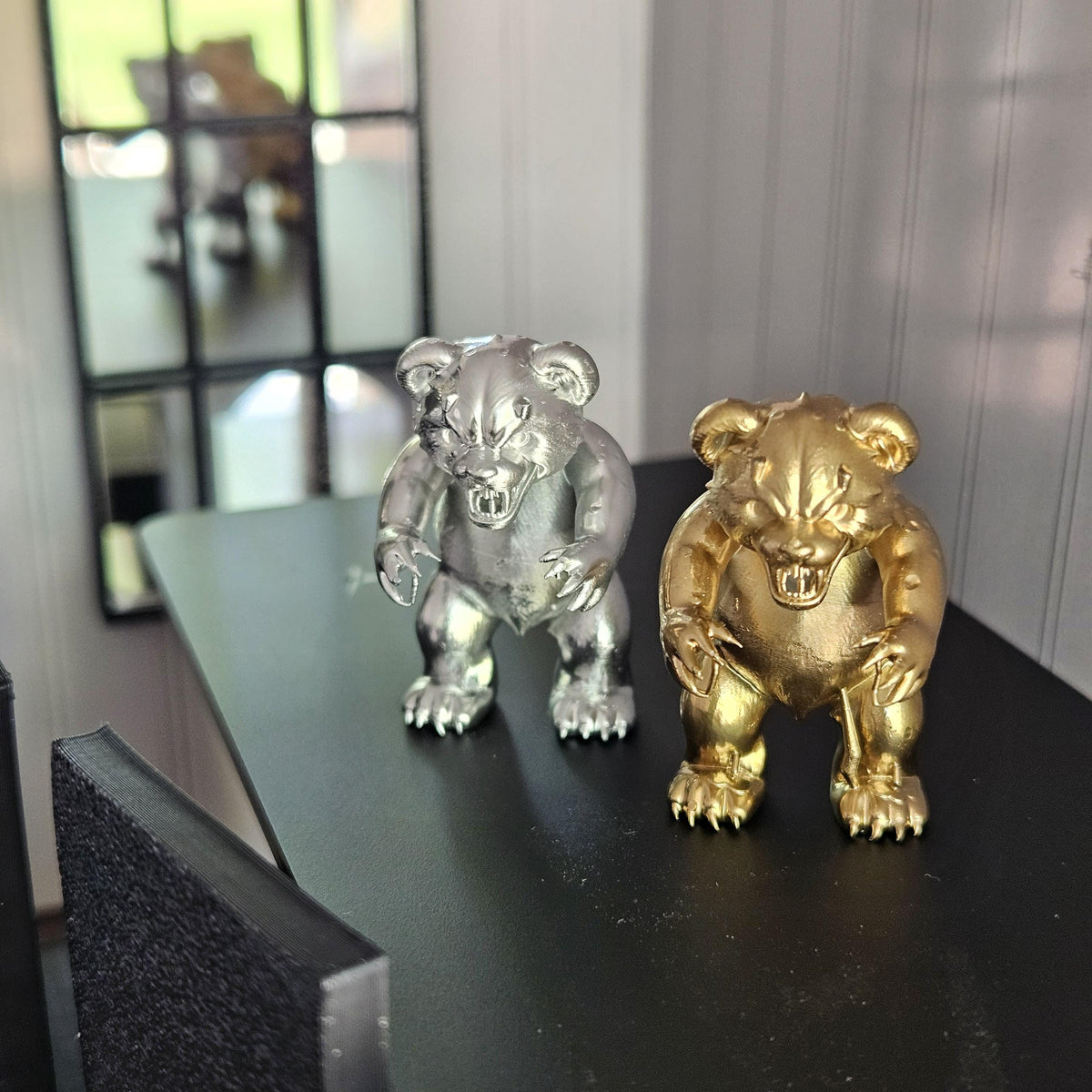 Silver and gold scary bear ornaments viewed slightly from above on a black table beside a miniature chair and mirror.