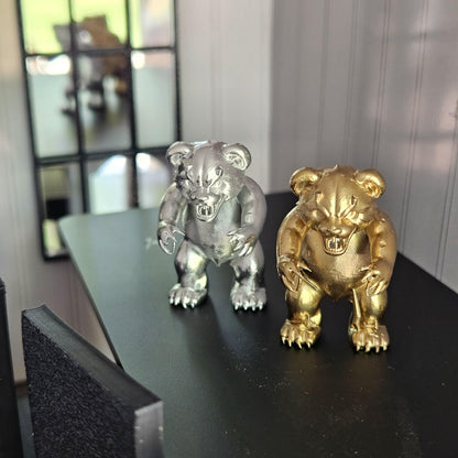 Silver and gold scary bear ornaments viewed slightly from above on a black table beside a miniature chair and mirror.