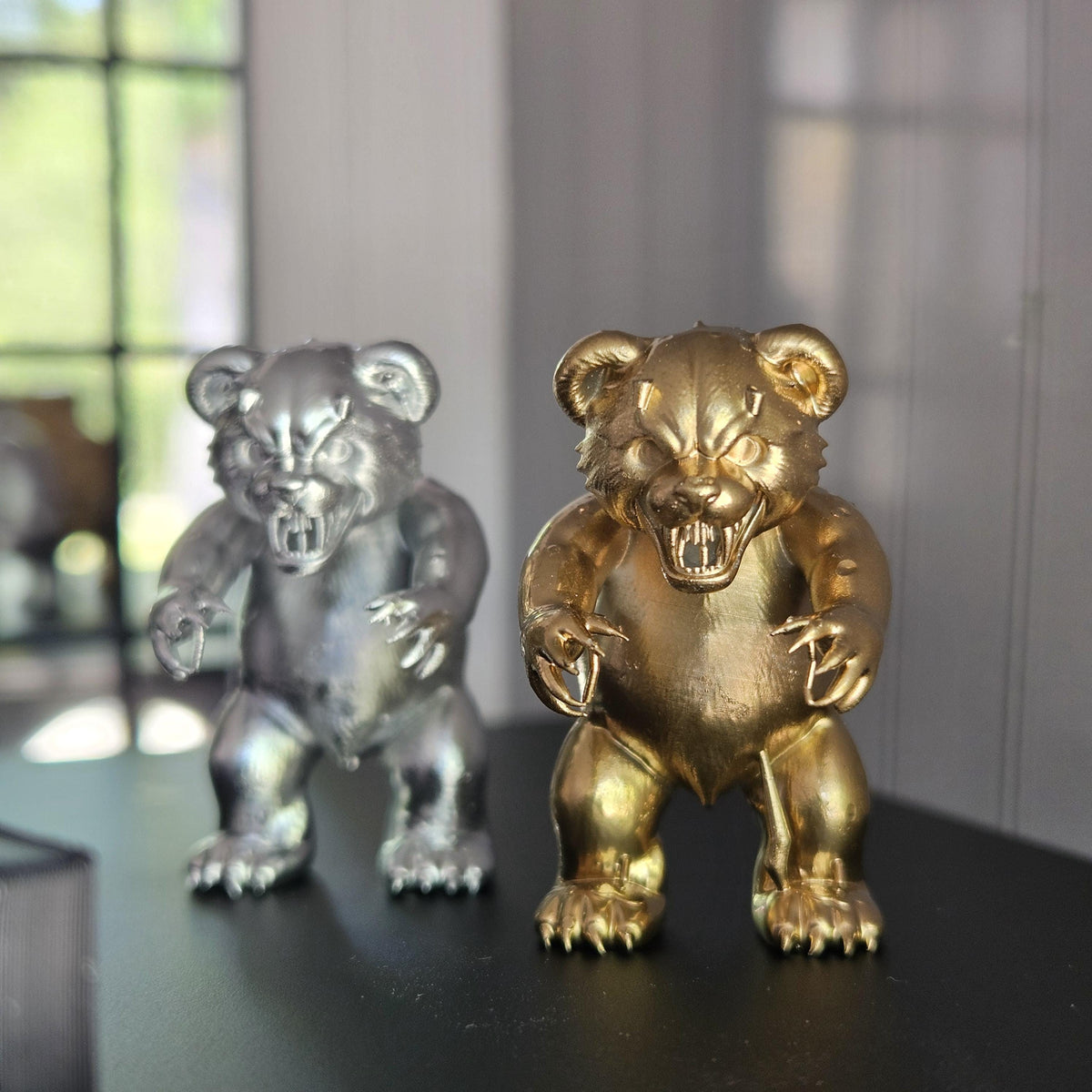Gold scary bear ornament in front of a silver version on a black tabletop, showing snarling faces and clawed paws.