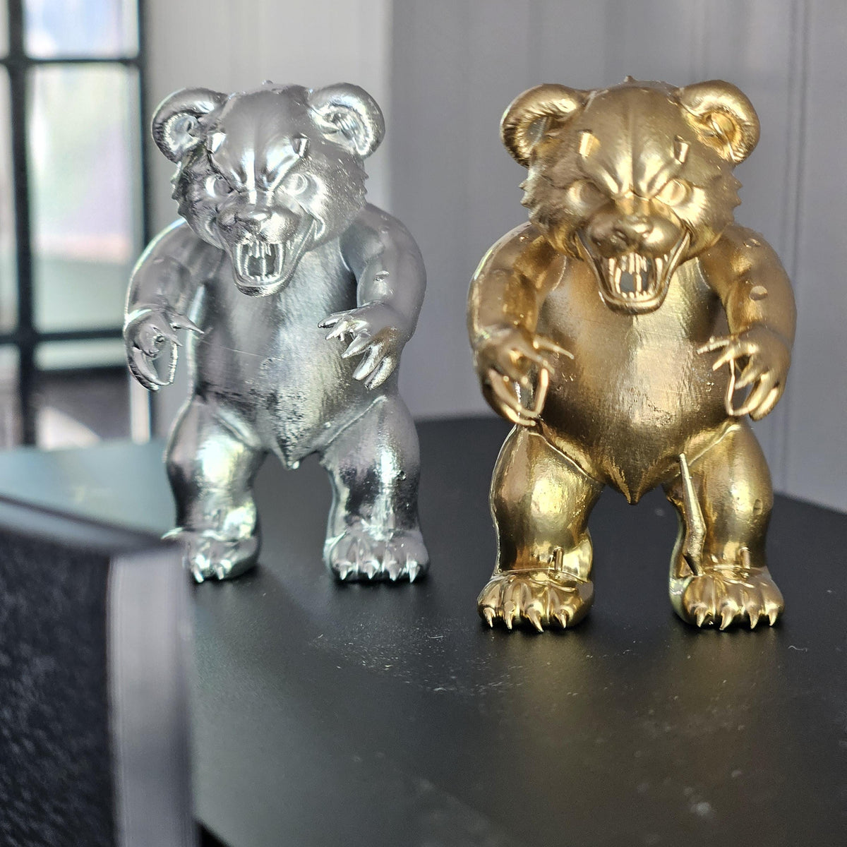 Front view of silver and gold scary bear ornaments side by side, showing rounded ears, bared teeth and clawed feet.