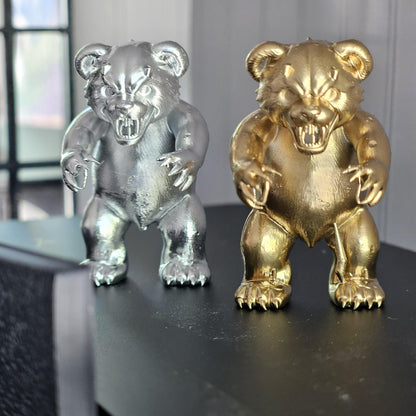 Front view of silver and gold scary bear ornaments side by side, showing rounded ears, bared teeth and clawed feet.
